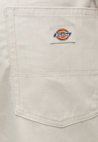 Τσέπη από μπεζ ύφασμα με ορατές ραφές και μικρό ορθογώνιο λογότυπο Dickies ραμμένο κοντά στο πάνω κέντρο.