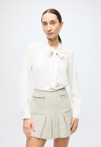 Witte blouse met lange mouwen en een striksluiting bij de kraag, gecombineerd met een lichtgekleurde, gestructureerde plissérok met zwarte accenten en zakken.