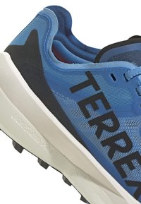 Scarpa sportiva blu con tomaia in rete, dettagli neri, logo "TERREX" ben visibile e suola bianca ammortizzata con tacchetti multidirezionali.