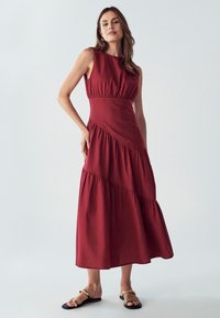 Rdeča, brezrokavna dolga obleka s prilegajočim zgornjim delom in plastičnim krilom. Narejena iz lahkega materiala, ki ima subtilno teksturo in ukrivljene šive.