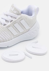 Bílá sportovní bota s texturovaným pleteným svrchním dílem, třemi bílými pruhy a polstrovanou podrážkou. Obsahuje dva páry bílých tkaniček.