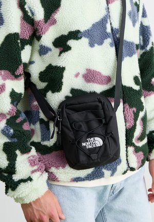 Osoba nosící vícebarevný fleecový kabát a světle modré džíny s malou černou crossbody taškou značky North Face.