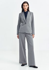 Szary dopasowany garnitur z marynarką i szerokimi spodniami, wykonany z fakturowanej tkaniny. Uzupełniony granatową koszulą z kołnierzykiem i czarnymi butami.