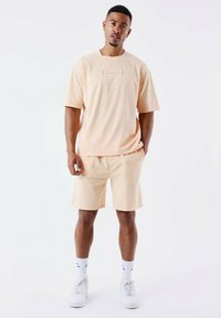 Camiseta de algodón de melocotón claro con cuello redondo sencillo y logotipo bordado, combinada con pantalones cortos a juego y zapatillas blancas, de pie sobre un fondo simple.