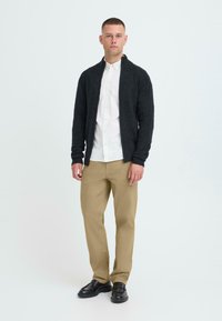 Grå lynlås-cardigan, hvid skjorte med knapper, beige bukser, sorte sko; glat tekstur, skræddersyet pasform og afslappet design.