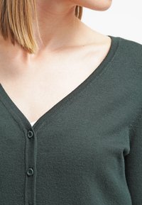 Mörkgrön cardigan med V-ringning, med en mjuk textur, två knappar fram och en slim fit, delvis visad på en modell.