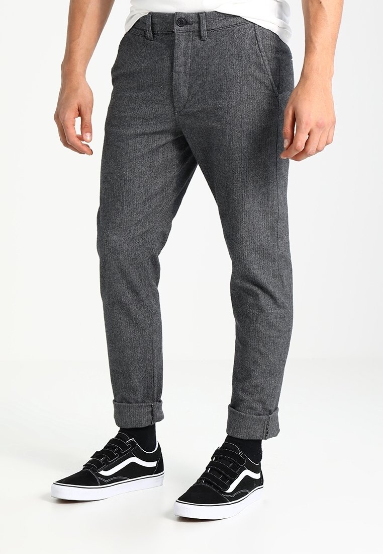 Grijze, taps toelopende broek in een structuurstof, met een knoopsluiting en opgerolde cuffs. Gecombineerd met zwarte sneakers en zwarte sokken.