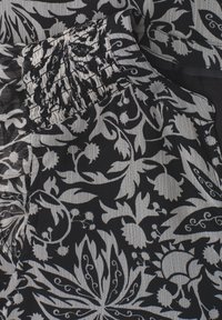 Tissu noir et blanc avec des motifs floraux et feuillus denses, présentant un détail de couture élastique froncée près du bord supérieur.