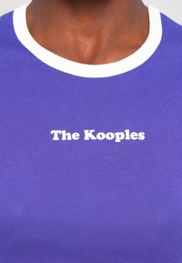 Short-sleeve purple T-shirt with a white collar; features white text reading "The Kooples" at the center. Smooth fabric texture.
