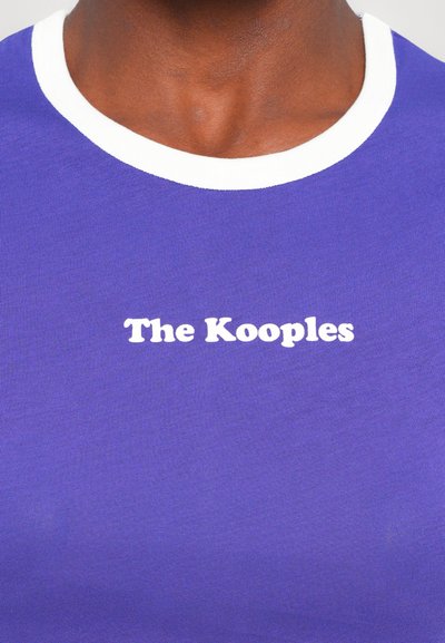 Īsu piedurkņu purpura T-krekls ar baltu apkakli; centrā ir balts uzraksts "The Kooples". Gluda auduma tekstūra.