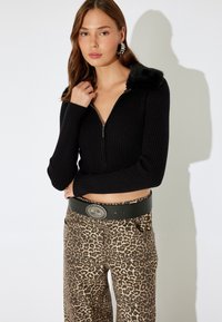 Schwarze gerippte Crop-Jacke mit Reißverschluss und Kragen aus Kunstpelz, kombiniert mit weit geschnittenen Hosen im Leopardenmuster und einem grünen Gürtel mit einer dekorativen Schnalle.