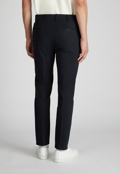 Pantaloni blu navy con vestibilità slim, realizzati in un tessuto liscio. Caratterizzati da tasche posteriori con chiusura a bottone e design davanti piatto. Indossati con sneakers bianche.