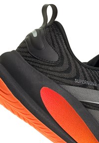 Talons de chaussure de sport noire avec semelle orange et détail rouge, dotée d'une languette de traction et du logo "SUPERNOVA" sur une tige en tissu texturé.