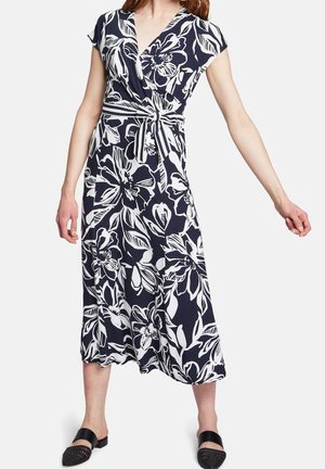 Mujer con vestido cruzado midi azul marino con estampado grande de flores blancas y cinturón de tela a rayas, combinado con zapatos negros sin cordones.