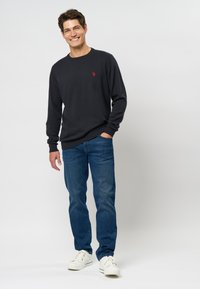 Marine crewneck trui, lange mouwen, geribbelde boorden. Luchtige blauwe jeans, witte sneakers, staand op een neutrale achtergrond met een glimlach.