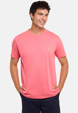 Joven sonriente con las manos en los bolsillos, lleva una camiseta rosa coral con un pequeño logo amarillo de una palmera y pantalones cortos oscuros.
