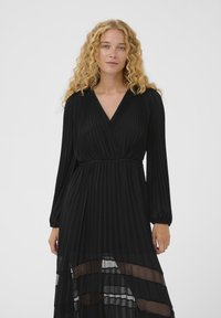 Vestido negro con plisados, escote en V profundo, mangas largas abullonadas y capas inferiores transparentes. Su textura suave y diseño en capas realzan su forma fluida.
