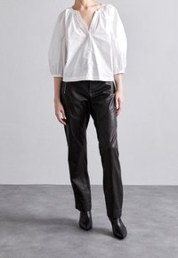 Weiße Baumwollbluse mit Puffärmeln und V-Ausschnitt, kombiniert mit schwarzen Lederhosen mit seitlichem Reißverschluss und figurbetontem Schnitt. Schwarze Stiefeletten mit Absatz.