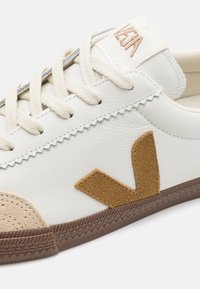 Hvite lær sneakers med beigefarget semsket tåhette, brun gummisåle, kremfargede snørebånd og en brun V-logo på siden, med stikkede detaljer.