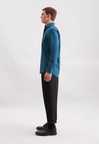 Mann steht seitlich und trägt ein türkisfarbenes Hemd mit Knöpfen, schwarze maßgeschneiderte Hosen, schwarze Socken und schwarze Dress-Schuhe vor einem weißen Hintergrund.