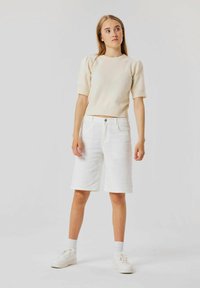 Krämfärgad stickad topp med kort ärm och vita denimshorts. Toppen har en rundad halsringning; shortsen är knälånga med fem fickor och en knapp.