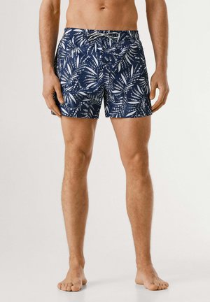 Modelo masculino vistiendo pantalones cortos de baño azul marino con patrones de hojas blancas, cintura elástica y cordón, de pie descalzo contra un fondo liso.