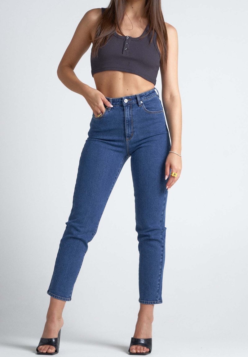 Femme portant un jean bleu taille haute, un débardeur court noir et des talons ouverts noirs, debout avec une main dans la poche sur un fond uni.
