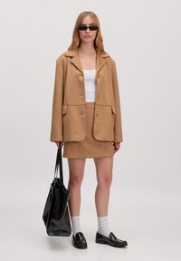 Beiger Blazer mit großen Revers und zwei frontalen Taschen, kombiniert mit einem passenden beigen Rock. Eine schwarze Tote-Bag und Sonnenbrille runden den Look ab.