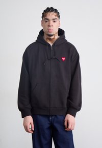 Black zip-up hoodie made of soft fabric, featuring a front pocket and a red heart logo on the left chest. Drawstring hood included.