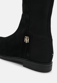 Bottine en daim noir avec un panneau latéral élastique côtelé, une semelle en caoutchouc plate et un petit logo doré sur le talon.