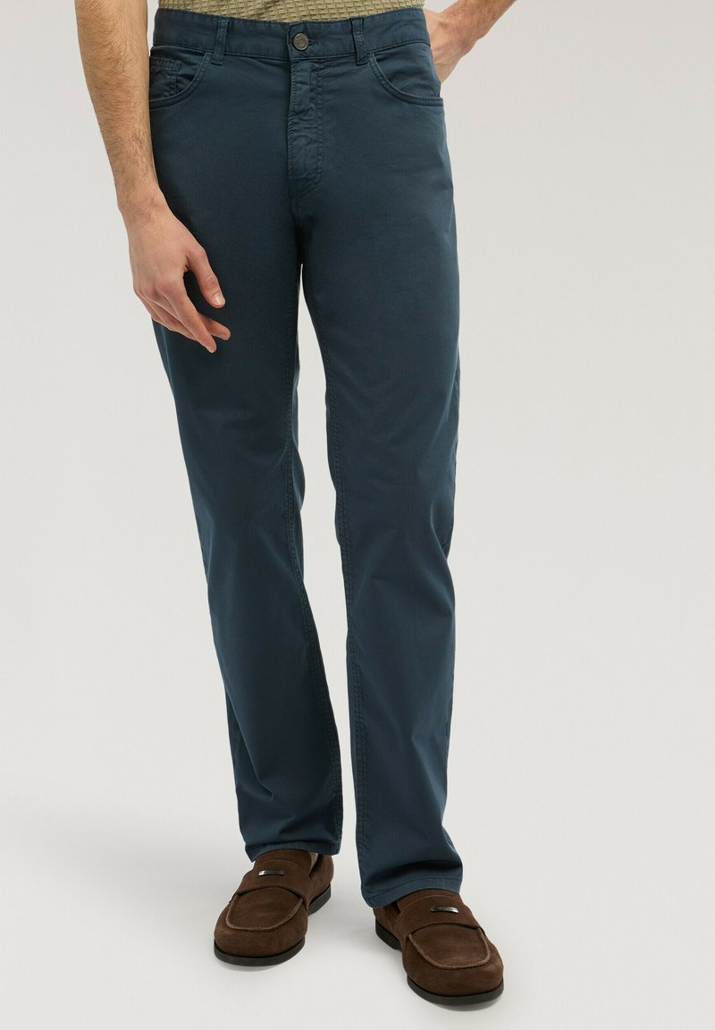 Uomo che indossa pantaloni dritti blu scuro e mocassini di camoscio marrone, in piedi con una mano visibile al suo fianco.