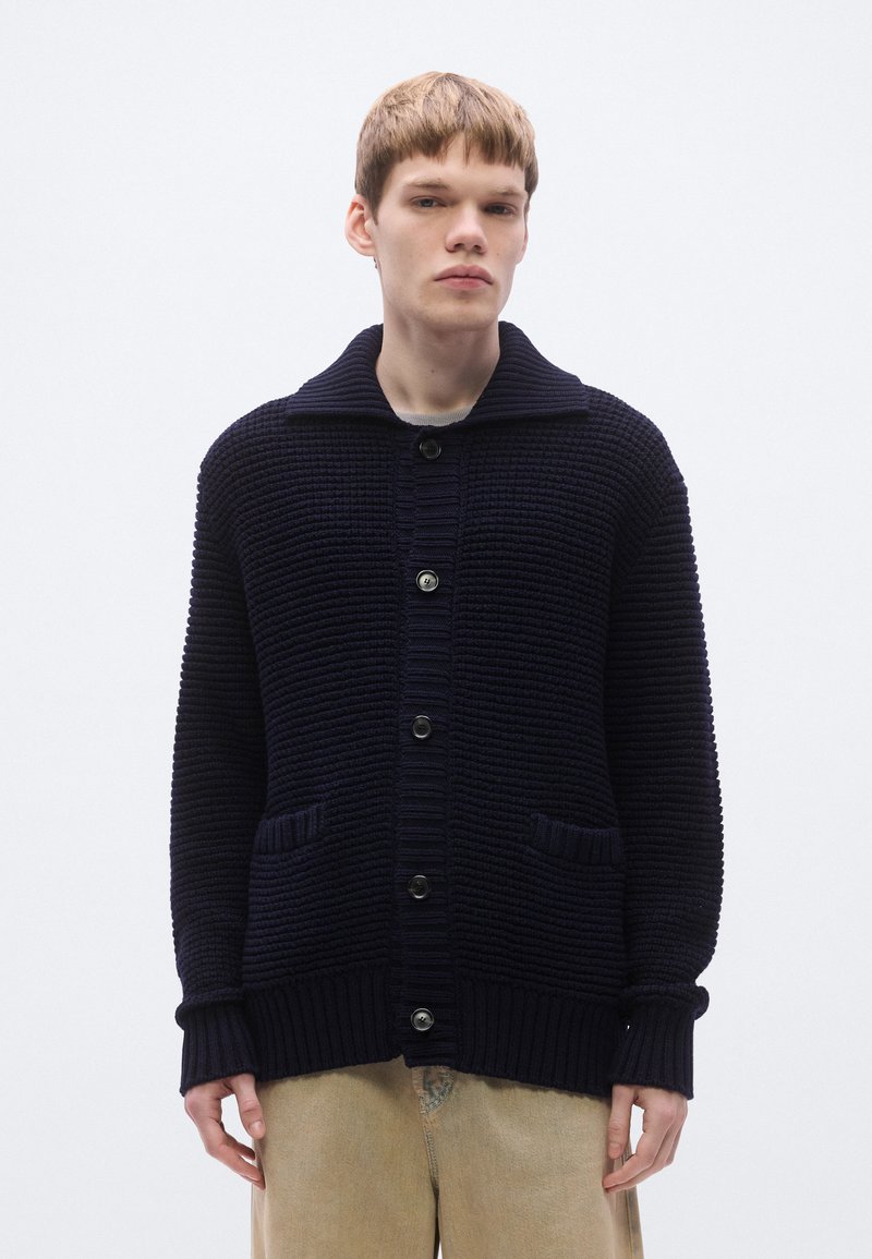 Tengerészkék kötött kardigán bordázott textúrával, nagy gallérral és három elülső gombbal. Két elülső zsebbel és bordázott mandzsettákkal rendelkezik.