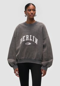 Sudadera gris de gran tamaño con "BERLÍN" en letras blancas en negrita. Presenta un cuello redondo y mangas voluminosas, confeccionada en una suave tela texturizada.