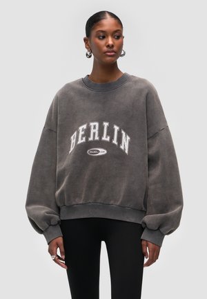 Grauer, übergroßer Sweatshirt mit "BERLIN" in fettem weißen Schriftzug. Mit Rundhalsausschnitt und voluminösen Ärmeln, aus weichem, texturiertem Stoff gefertigt.