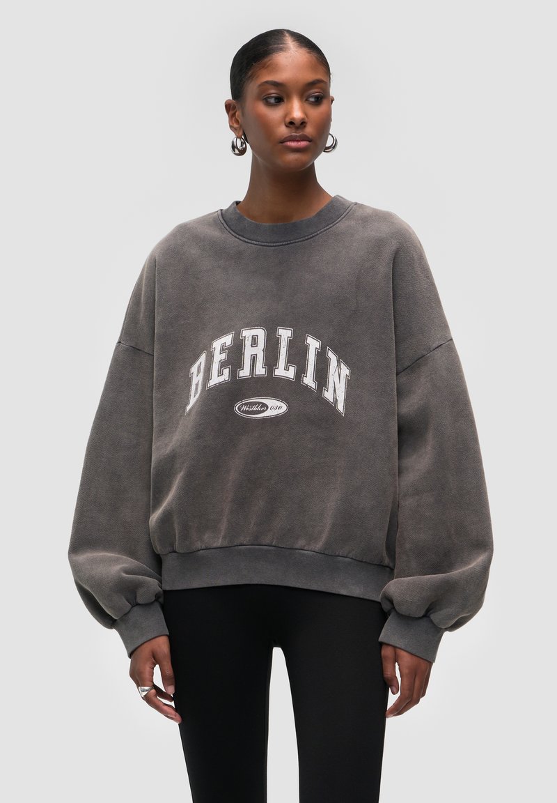 Sudadera gris de gran tamaño con "BERLÍN" en letras blancas en negrita. Presenta un cuello redondo y mangas voluminosas, confeccionada en una suave tela texturizada.