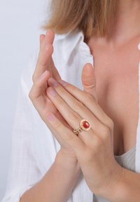 Bague en or avec une pierre ovale rouge, pr ésentant un design de bande tressée. Des mains sont jointes, mettant en avant les détails de la bague.