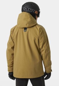 Giacca da sci impermeabile marrone con cappuccio, polsini elasticizzati e etichetta con marchio sul retro; indossata con guanti neri e un casco nero.