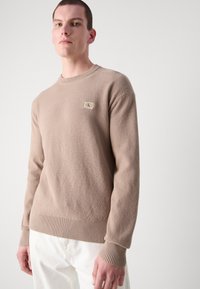 Maglione beige a maglia con un motivo testurizzato, polsini e orlo a costine, con una piccola toppa con logo sul petto. Il modello indossa pantaloni chiari.