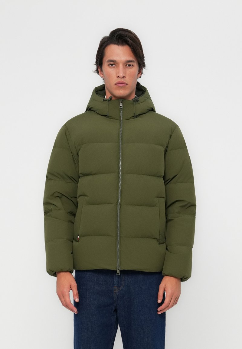 Jachetă puffer de culoare verde măslinie, cu glugă, design căptușit, închidere cu fermoar frontal și buzunare laterale, purtată cu blugi albaștri.