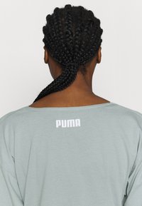 Vaaleanharmaa ylisuurikokoinen t-paita, jossa on leveä kaula-aukko, pehmeä rakenne ja valkoinen "PUMA"-logo painettuna yläselkään.