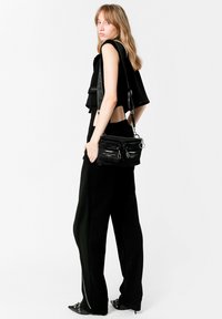 Bolso negro de hombro con textura de tela, detalles de cremalleras y herrajes metálicos, combinado con un top negro corto y pantalones negros de pierna ancha.