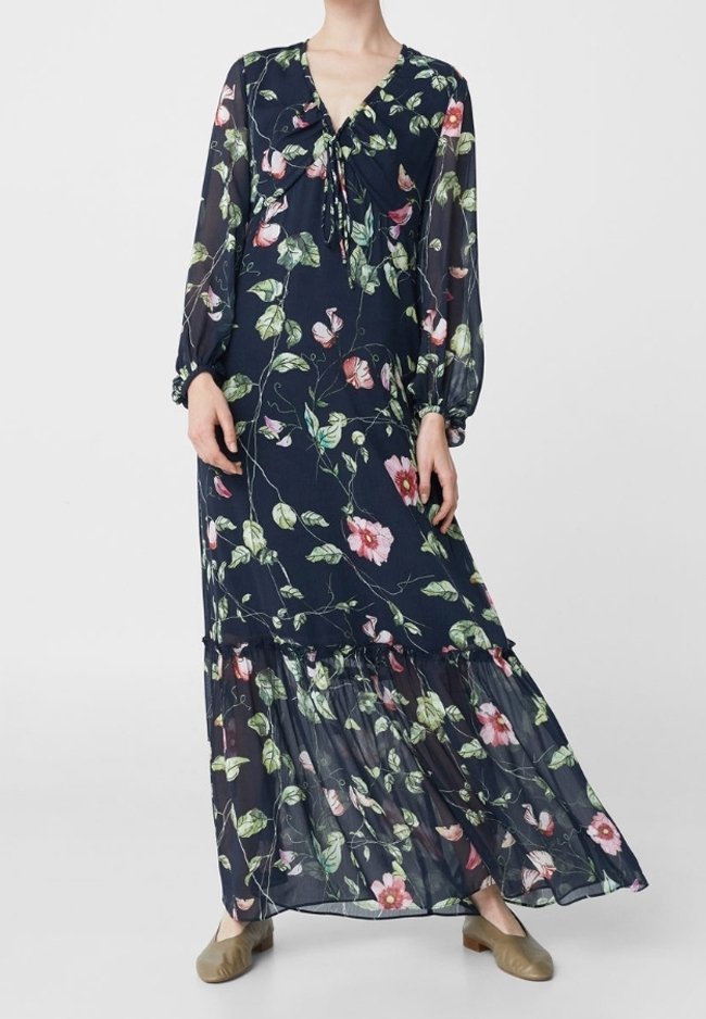 Robe maxi bleu marine à imprimé floral rose, manches longues transparentes, ourlet à volants et col avec cordon, portée avec des ballerines beige.