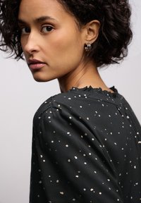 Musta paita hopeisilla pilkuilla, jossa on rypytetty kaula-aukko ja poimutettu olka. Sileä pinta ja rento istuvuus.