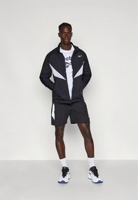 Must spordijakk valgete detailidega, sellega on kombineeritud valge logoga t-särk ja mustad lühikesed püksid. Jalanõudel on mustad paelad ja valged tallad.