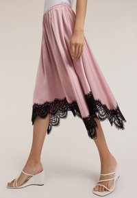 Gonna di raso rosa con una vita elastica, caratterizzata da un orlo in pizzo nero e una silhouette fluida. Indossata con sandali a zeppa bianchi.