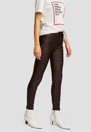 Kvinde iført hvid t-shirt med grafik, mørkebrune tætsiddende bukser og hvide ankelstøvler med hæle, stående mod en ensfarvet baggrund.