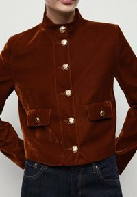 Giacca in velluto marrone con colletto alto, bottoni dorati e due tasche frontali. Presenta un design aderente e una superficie liscia.