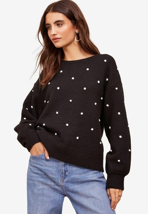 Black knitted sweater featuring white polka dots. Loose fit with a rounded neckline and ribbed cuffs. Worn with light blue jeans.