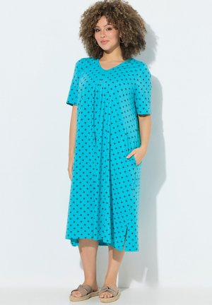 Kvinde med krøllet hår iført en blå midi-kjole med sorte stjernemønstre og beige sandaler, står med den ene hånd i kjolens lomme.