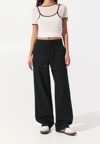 White short-sleeve top with navy trim, paired with loose black pants featuring an elastic waistband and gold hardware accents. Casual sneakers.